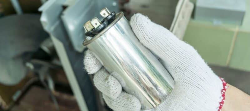 Close-up of a gloved hand holding a cylindrical capacitor, typically used in HVAC systems. The background shows blurred machinery and equipment, indicating a maintenance or repair setting